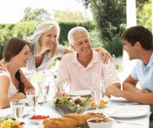 A group of four people, two men and two women, are sitting at an outdoor table, smiling and chatting. The table is set with glasses, plates, and a salad bowl. They are surrounded by greenery on a sunny day.