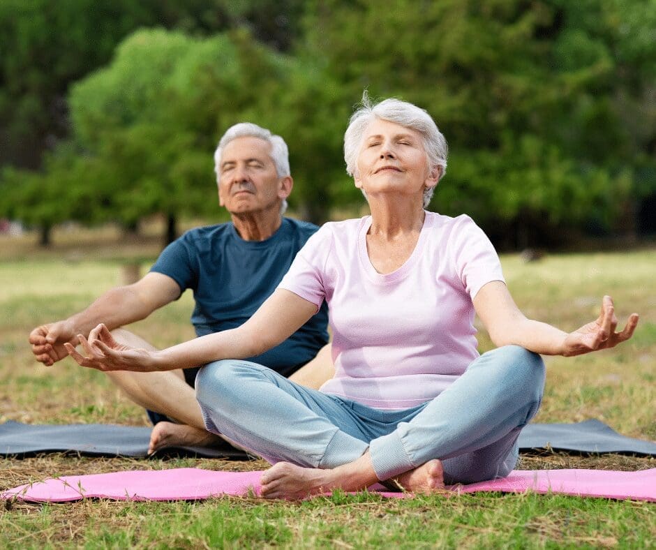 Elderly man and woman practicing yoga outdoors on mats in a park. Both are seated in a cross-legged position with eyes closed, appearing relaxed and serene. Trees and grass are visible in the background.