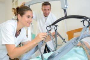 Two healthcare professionals in white uniforms adjust a patient lift over a hospital bed. The female is focused on attaching straps to the lift, while her male colleague observes closely. The room, equipped with various medical devices, embodies the precision and care essential in modern healthcare.