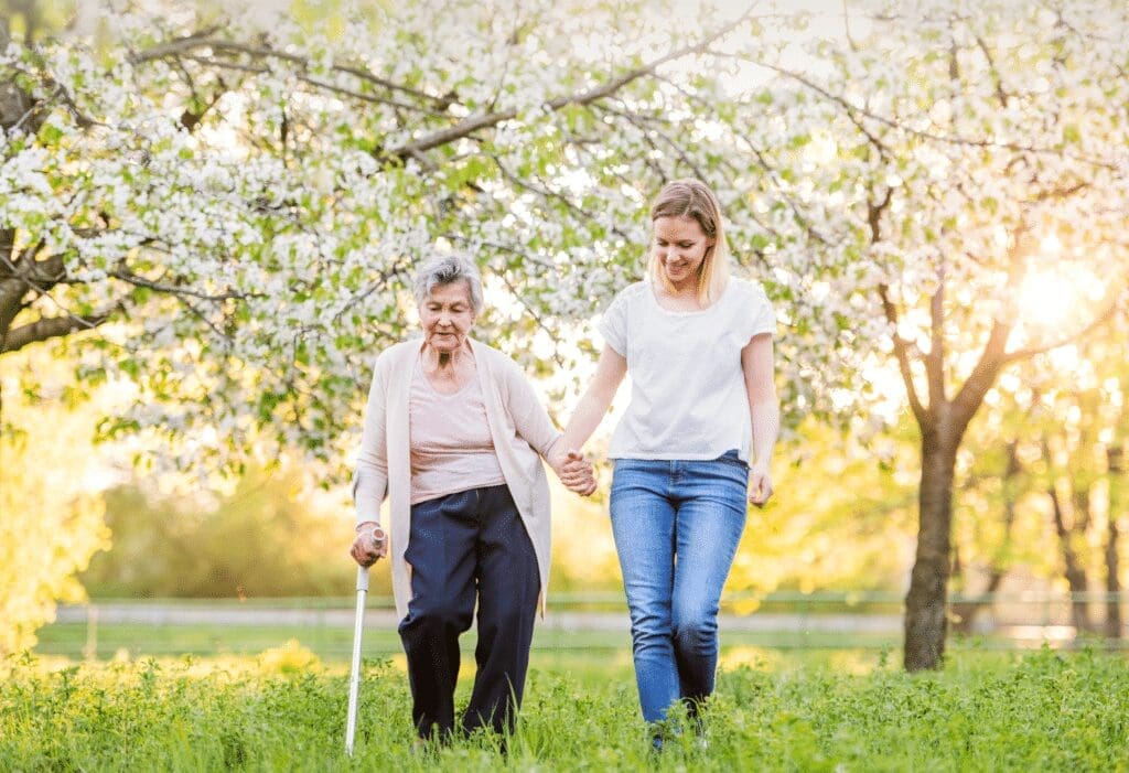 A young woman and an elderly woman, with a rollator by her side, walk hand in hand through a grassy area with blooming trees in the background. The sun is shining, casting a warm glow over the scene.