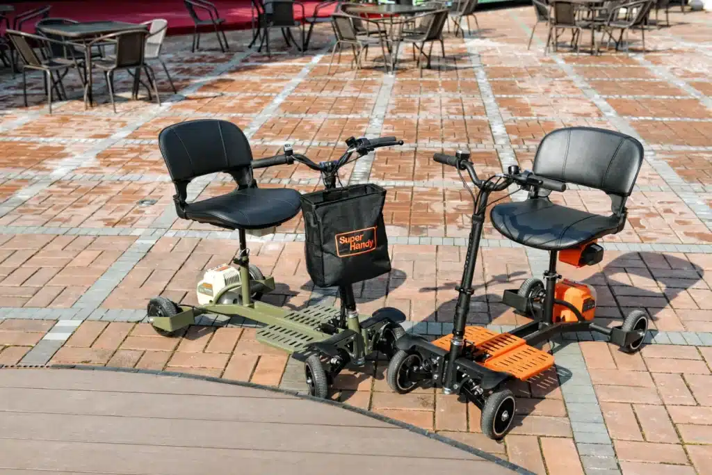 Two scooters, including a SuperHandy Passport Pro, are parked outdoors on brick. One is green and the other orange, both having black seats, handlebars, and accessory pouches. Tables and chairs appear in the background.
