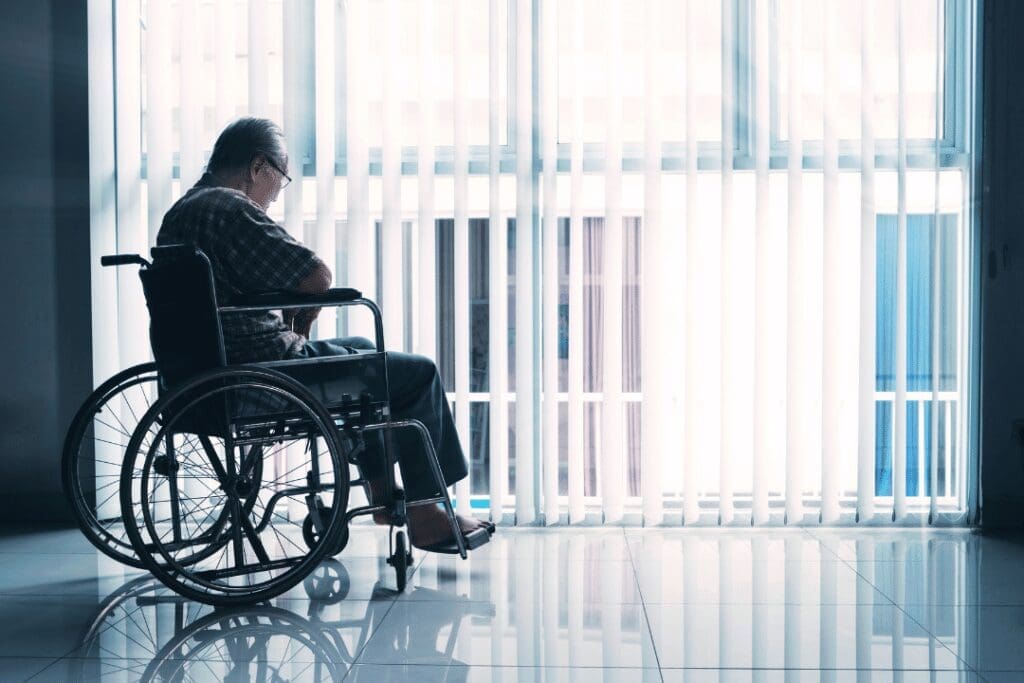 An elderly person in a wheelchair sits near a large window with vertical blinds, gazing outside. The room is softly illuminated by natural light, casting reflections on the polished floor.