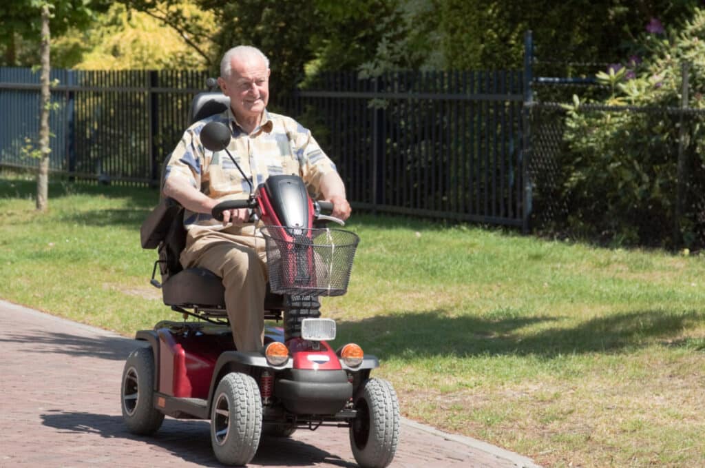 An elderly man rides a mobility scooter on a sunny day, following a paved path in a park. He is wearing a short-sleeve shirt and khaki pants. Green grass and trees are visible in the background, with a black fence on one side.