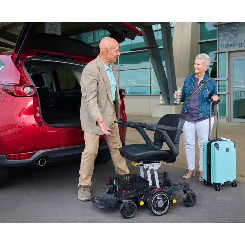 An older couple stands by a red car before a building. The man points to a Buzzaround Carry On Powerchair, while the woman holds a water bottle and blue suitcase, suggesting they are at a travel hub.