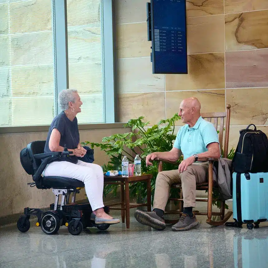 Two older adults chat indoors; one sits in a Buzzaround Carry On Powerchair, the other in a rocking chair. A blue suitcase and backpack lie near a small table and large green plant, creating a cozy atmosphere.