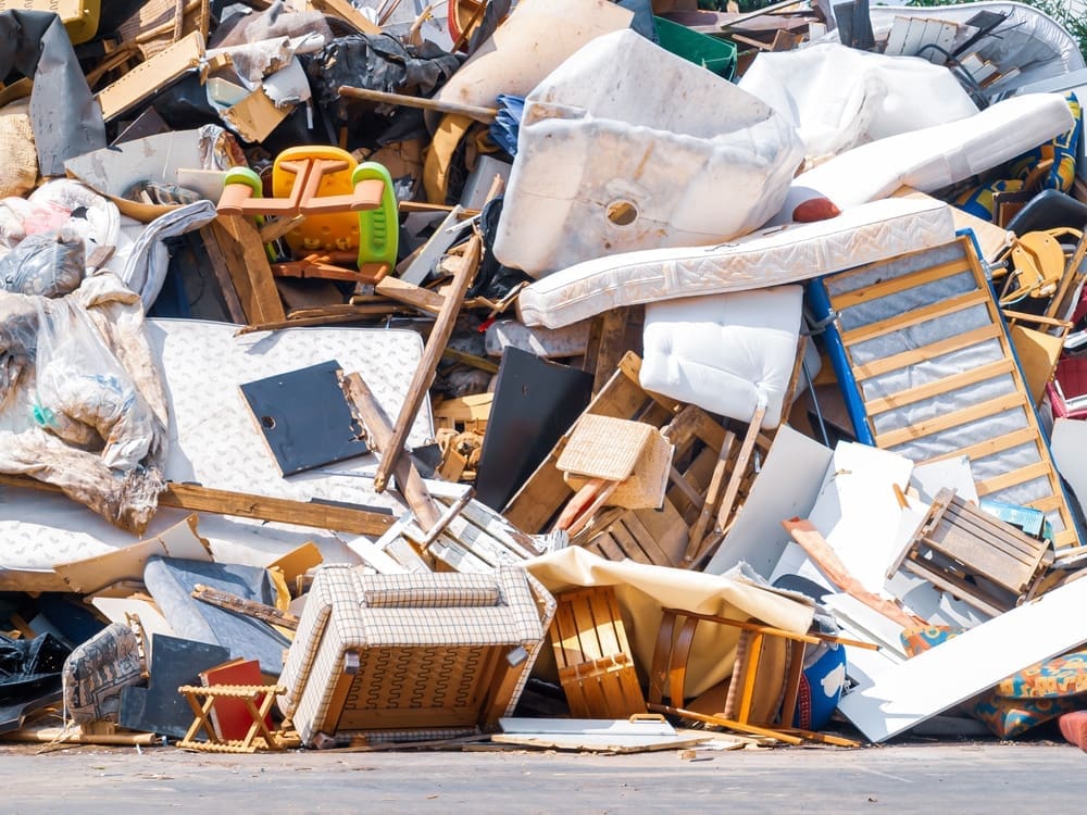 A heap of discarded furniture and household items in a landfill. The pile includes mattresses, chairs, wooden pieces, and a childs plastic seat amongst other debris. The items appear worn and are haphazardly stacked.
