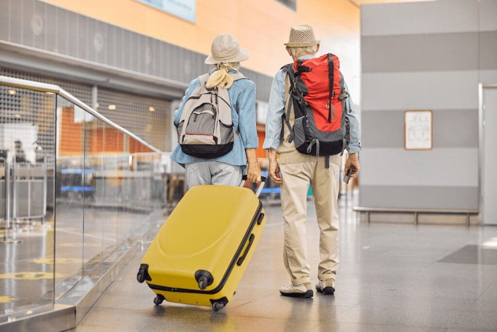 Two older adults in casual travel attire walk through an airport with backpacks and a large yellow suitcase. The setting has modern architecture with metal railings and a patterned floor. They appear to be engaged and focused on their journey.