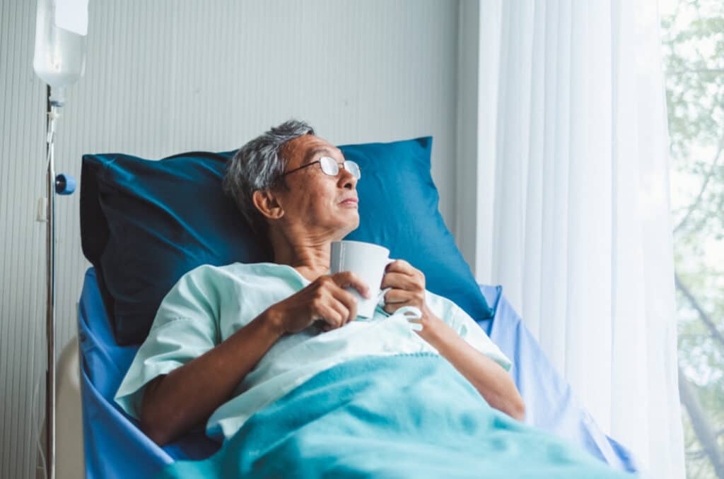 An elderly person with glasses lies in a hospital bed, holding a white mug and looking out a window. They are covered with a turquoise blanket, and theres an IV pole next to the bed. Sunlight filters through a curtain.