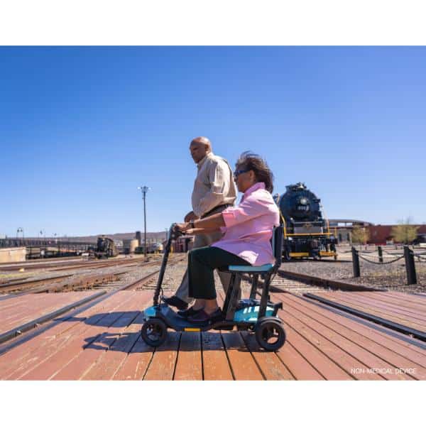 An elderly woman rides a Go Go Super Portable on a wooden boardwalk with an elderly man walking beside her, while a vintage train is visible against the clear blue sky in the background.