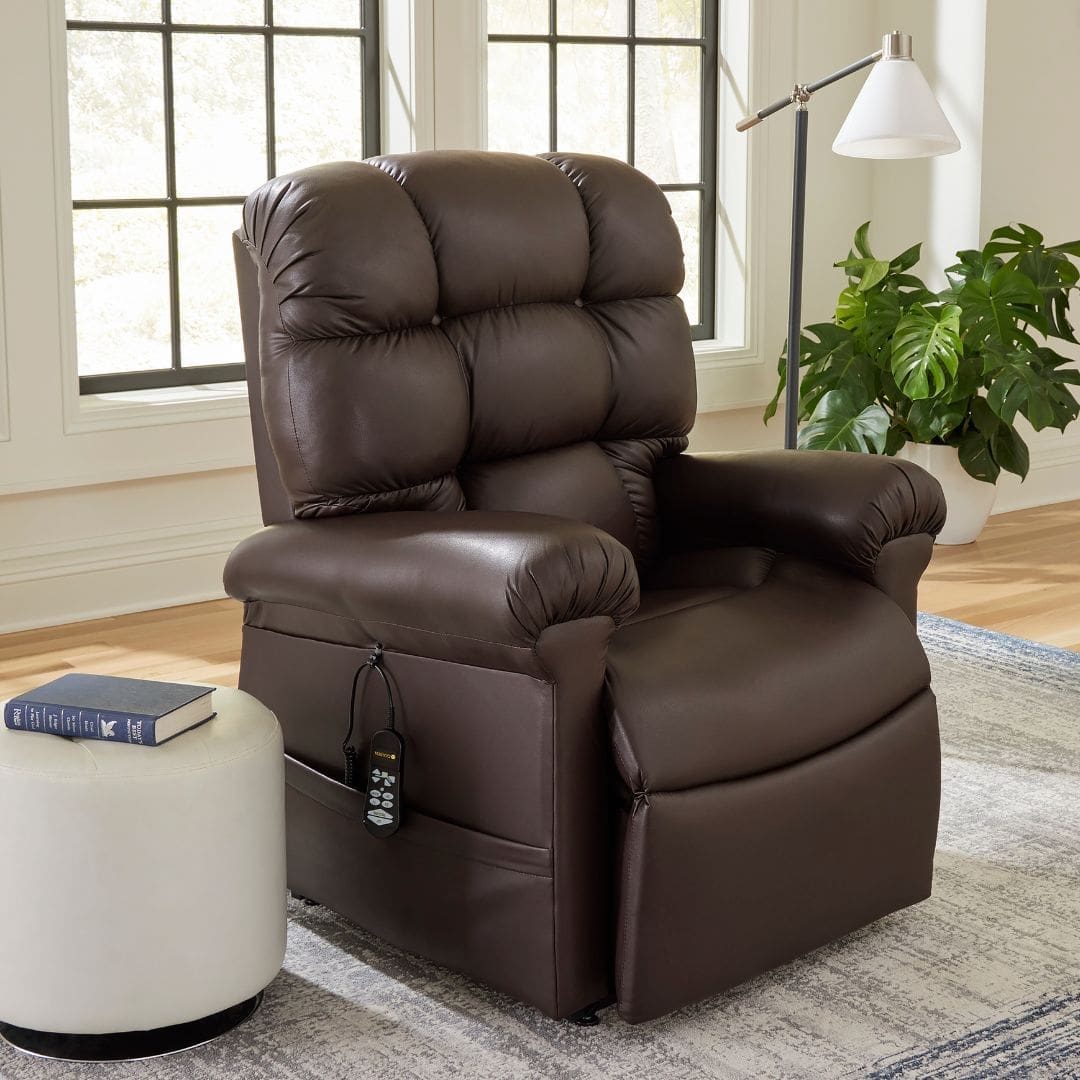 In a bright room with large windows, the luxurious Golden Cloud PR 517 recliner with Power Headrest & Lumbar sits beside a white floor lamp and a large green plant in the corner, while a round white ottoman nearby holds a book.