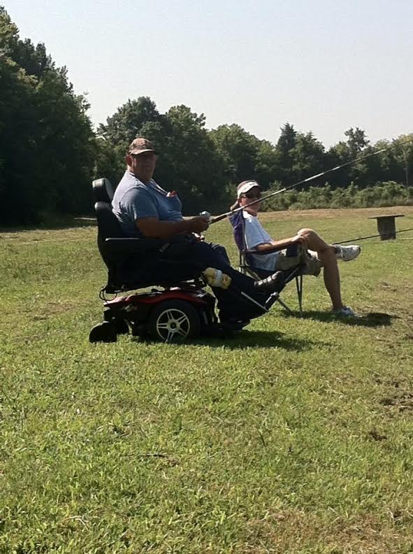 Two people are fishing in a grassy area. One person is sitting in a motorized wheelchair, while the other sits in a folding chair. Both are holding fishing rods. Trees and clear skies are in the background.