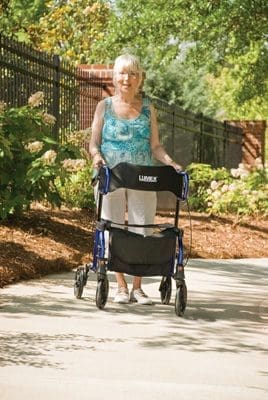 An elderly woman stands on a pathway in a garden, holding a walker. Shes wearing a blue-green sleeveless top, white pants, and white shoes. Lush greenery and a black fence are in the background under a sunny sky.